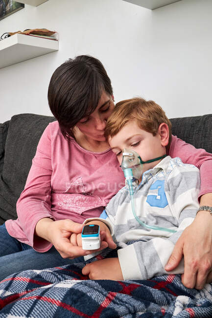 Mulher carinhosa usando oxímetro de pulso no dedo do menino doente em máscara de oxigênio usando nebulizador durante a inalação em casa — Fotografia de Stock