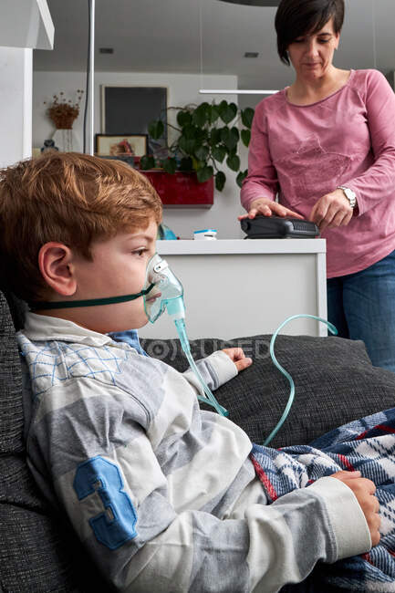 High angle of crop mother using nebulizer for inhalation of boy sitting in oxygen mask on sofa — Stock Photo