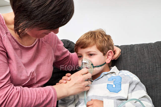 Contenido madre que se pone la máscara de oxígeno en la cara del hijo para el procedimiento de inhalación mientras está sentado en el sofá en casa - foto de stock