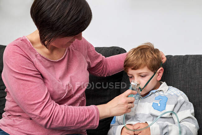 Conteúdo mãe colocando máscara de oxigênio no rosto do filho para procedimento de inalação enquanto se senta no sofá em casa — Fotografia de Stock