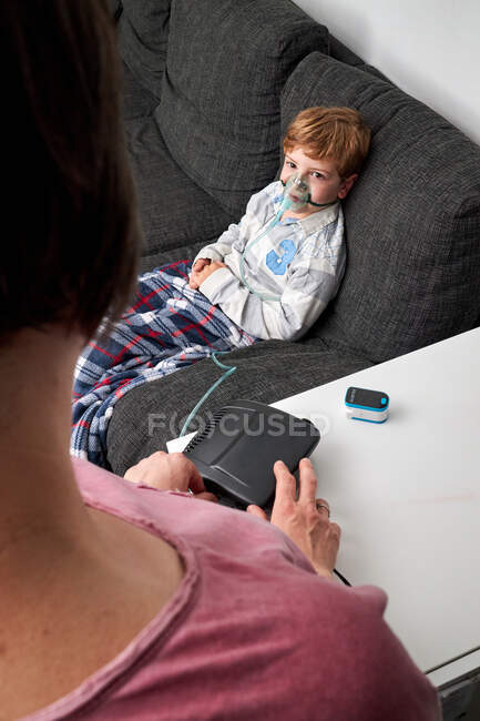 Ângulo alto de mãe de colheita usando nebulizador para inalação de menino sentado em máscara de oxigênio no sofá — Fotografia de Stock