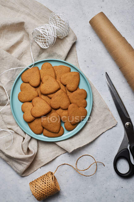 Vista superior del diseño de galletas de Navidad en forma de corazón dulce en el plato y materiales de embalaje surtidos en la mesa - foto de stock