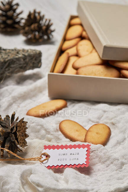 High angle of gift tag and cones placed on table with box full of homemade sweet Christmas biscuits — Stock Photo