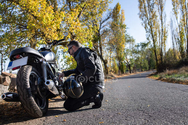 Vista lateral do piloto masculino maduro em jaqueta de couro ajoelhado e verificando o motor de bicicleta durante a viagem de outono na estrada de asfalto no campo — Fotografia de Stock