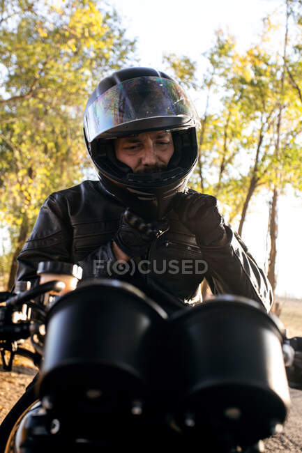 Концентрированный мужчина-гонщик в кожаной куртке, закрепляющий шлем и сидящий на мотоцикле осенью солнечного дня — стоковое фото