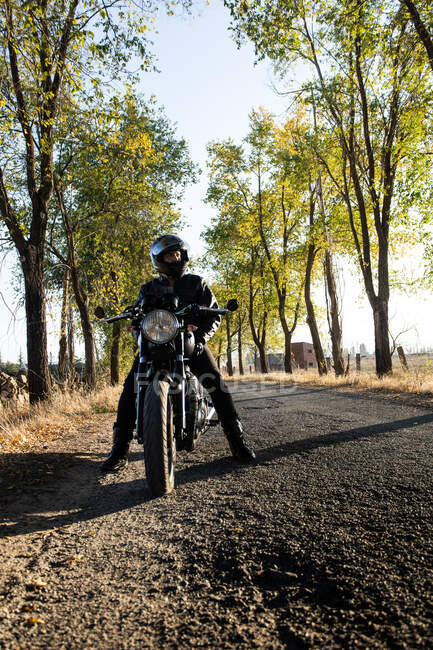 Motociclista irreconocible en casco y chaqueta de cuero sentado en motocicleta durante el viaje de aventura en el campo - foto de stock