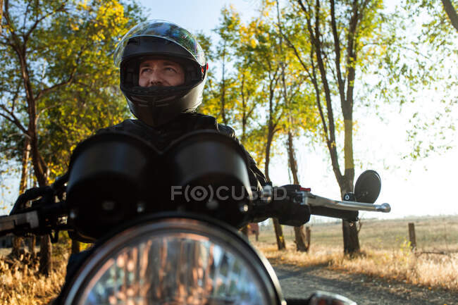 Racer masculino envejecido concentrado en chaqueta de cuero fijación casco y sentado en moto en el día soleado otoño - foto de stock
