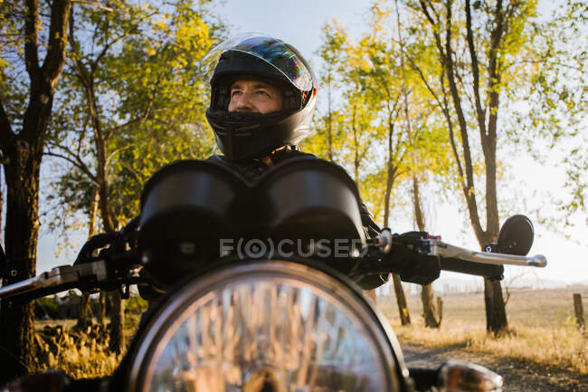 Концентрированный мужчина-гонщик в кожаной куртке, закрепляющий шлем и сидящий на мотоцикле осенью солнечного дня — стоковое фото