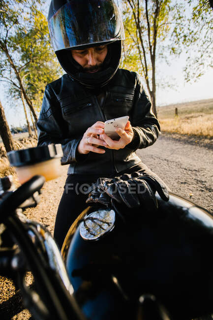 Концентрированный мужчина гонщик в кожаной куртке сидит на мотоцикле и просматривает телефон в осенний солнечный день — стоковое фото