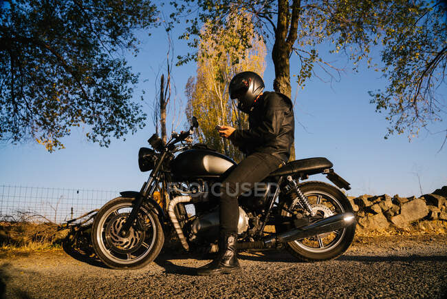 Сконцентрований літній чоловічий гонщик у шкіряній куртці кріпиться до шолома і сидить на мотоциклі восени сонячного дня. — стокове фото