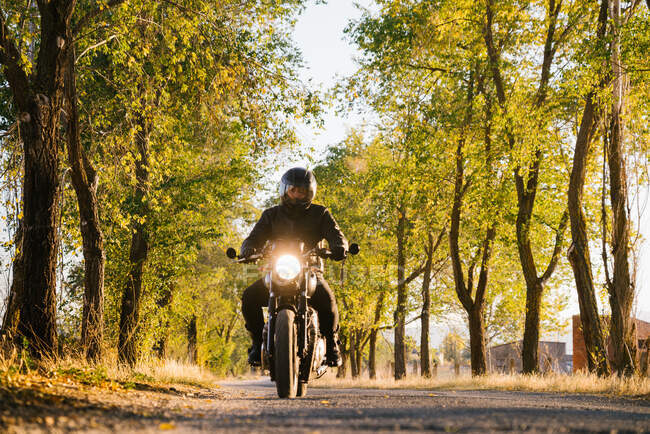 Hombre en chaqueta de cuero y casco bicicleta de montar en asfalto carretera en el soleado día de otoño en el campo - foto de stock