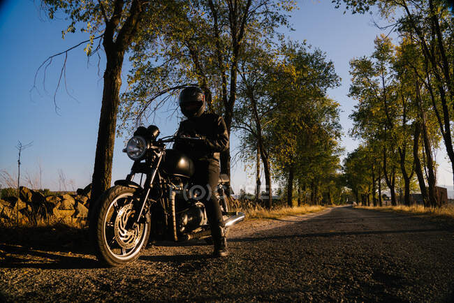 Концентрированный мужчина гонщик в кожаной куртке сидит на мотоцикле и просматривает телефон в осенний солнечный день — стоковое фото