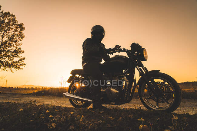 Motociclista sin rostro en casco a caballo motocicleta en la puesta del sol por la noche en la carretera rural - foto de stock