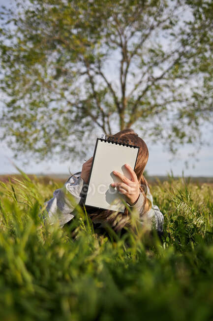 Menina adolescente irreconhecível sentado no prado e desenho em caderno de esboços desfrutando de dia ensolarado no campo — Fotografia de Stock