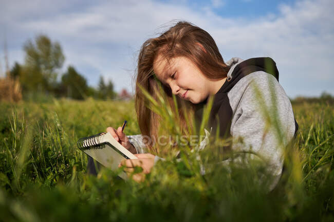 Entzückte Teenie-Mädchen sitzen auf der Wiese und zeichnen in Skizzenbuch, während sie sonnigen Tag in der Natur genießen — Stockfoto
