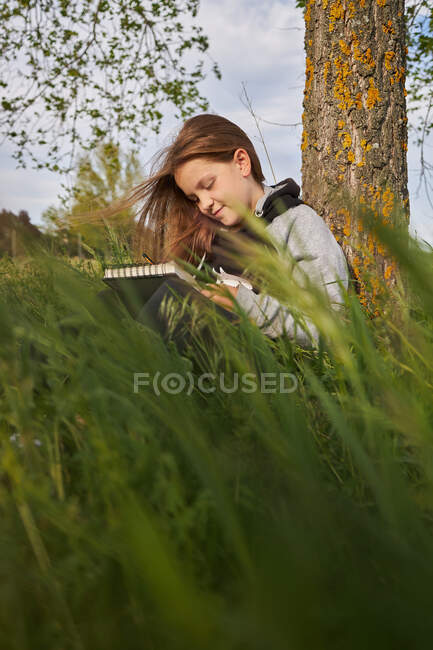 Delighted teen girl sitting in meadow and drawing in sketchbook while enjoying sunny day in countryside leaning on tree trunk — Stock Photo