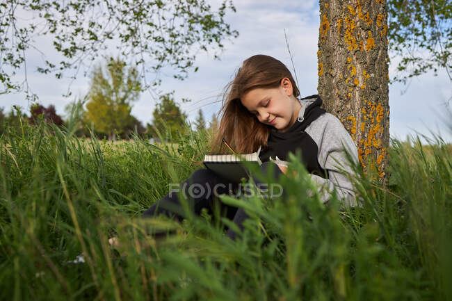 Side view of delighted teen girl sitting in meadow and drawing in sketchbook while enjoying sunny day in countryside leaning on tree trunk — Stock Photo