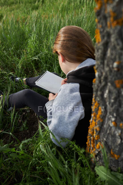 Rückansicht des nicht wiederzuerkennenden Teenie-Mädchens, das auf der Wiese sitzt und im Skizzenbuch zeichnet, während es den sonnigen Tag in der Natur genießt, angelehnt an einen Baumstamm — Stockfoto