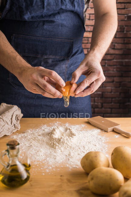 Uomo irriconoscibile in grembiule che rompe il tuorlo d'uovo in mucchio di farina di frumento vicino alle patate e olio durante la preparazione dell'impasto degli gnocchi sul tavolo in cucina — Foto stock