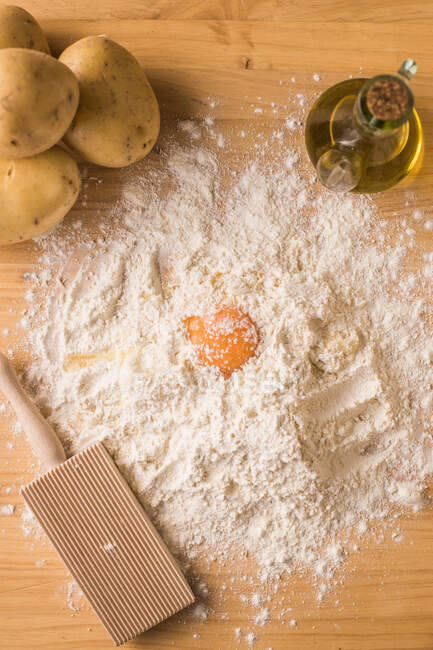 De arriba la yema de huevo puesta al montón de la harina de trigo cerca de las patatas y el aceite durante los ñoquis testo la preparación a la mesa en la cocina - foto de stock