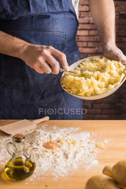 Hombre irreconocible en delantal añadiendo puré de papas a la harina y huevo crudo mientras prepara masa para ñoquis en la cocina - foto de stock