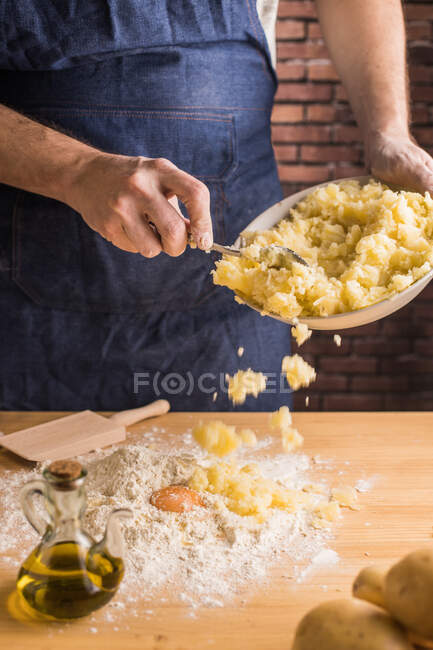 Невпізнаваний чоловік в фартусі додає картопляне пюре до борошна і сире яйце під час приготування тіста для ноккі на кухні — стокове фото