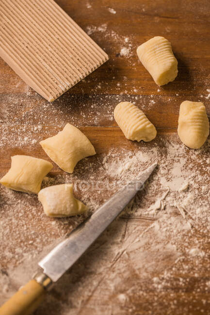 Vue de dessus des morceaux de pâte molle crue placés sur une table en bois recouverte de farine près du panneau de nervure et du couteau pendant la préparation des gnocchis dans la cuisine — Photo de stock