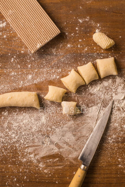 Vista dall'alto di pezzetti di pasta morbida cruda appoggiati su tavolo di legno ricoperto di farina vicino al tagliere e coltello durante la preparazione degli gnocchi in cucina — Foto stock