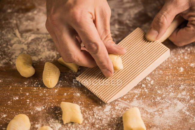 Неузнаваемые руки повара прижимают кусок теста к ребристой доске во время приготовления ньокки на деревянном столе, покрытом мукой — стоковое фото