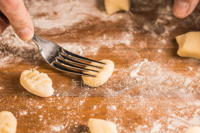 Unrecognizable person pressing small piece of dough with fork while cooking gnocchi on lumber table covered with flour at home — Stock Photo