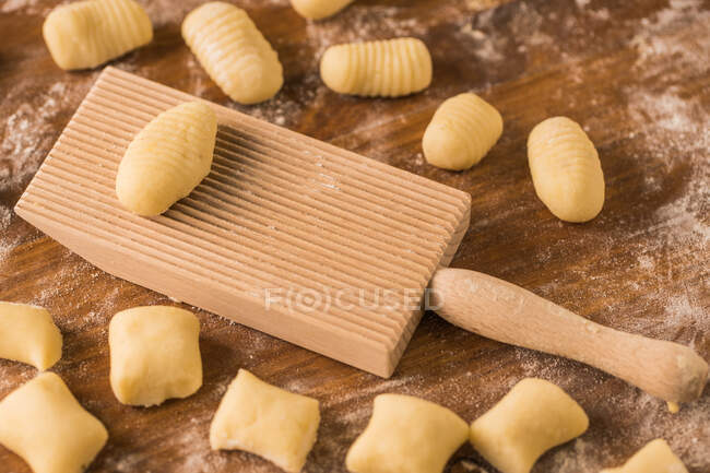 Vue de dessus des morceaux de pâte molle crue placés sur une table en bois recouverte de farine près du panneau de nervure pendant la préparation des gnocchis dans la cuisine — Photo de stock