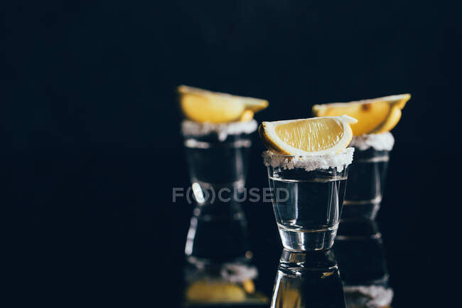 Colpi di tequila con sale e limone posti su una superficie riflettente su sfondo scuro — Foto stock