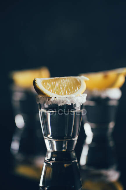 Tequila tiros com sal e limão colocados na superfície reflexiva contra fundo escuro — Fotografia de Stock