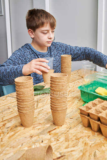 Niño sentado en la mesa con un montón de tazas de cartón y recipientes contra la pala de jardinería y tenedor mientras mira hacia otro lado - foto de stock
