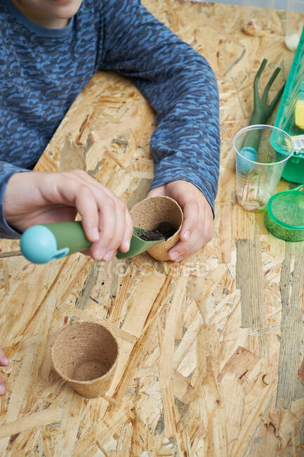 From above of crop anonymous kid with gardening shovel filling eco cup with earth at table — Stock Photo
