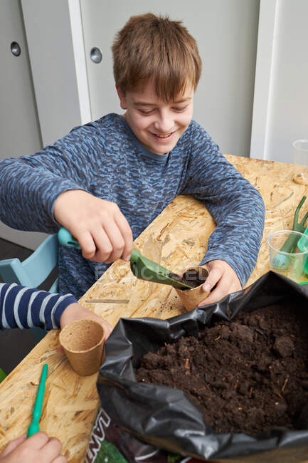 Niño feliz con jardinería pala relleno eco taza con tierra en la mesa - foto de stock