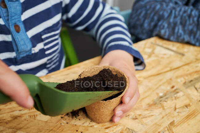 Anonymes Kind mit Gartenschaufel befüllen Ökobecher mit Erde am Tisch — Stockfoto