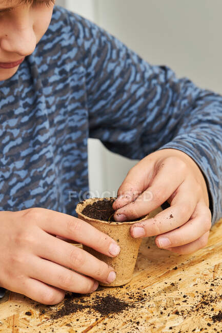 Cultivo niño anónimo plantación de plántulas en taza de cartón con tierra en la mesa con pala de jardinería - foto de stock