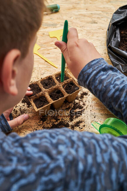 Зверху рослин анонімний дитина з насіннєвим дозатором копає отвори в біорозкладаній ємності з грунтом — стокове фото