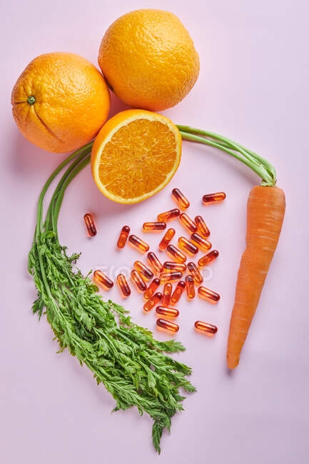 Par dessus la composition de pilules de vitamines dispersées disposées sur une table rose près de carottes mûres et d'oranges juteuses — Photo de stock