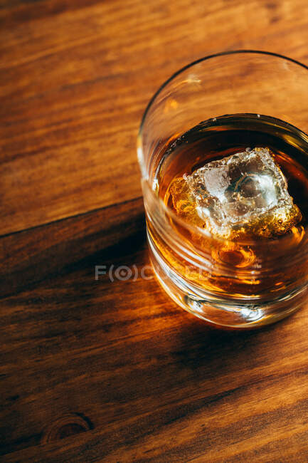 Зверху скляна чашка з холодним віскі та кубиком льоду, розміщена на столі з пиломатеріалів у темній кімнаті — стокове фото