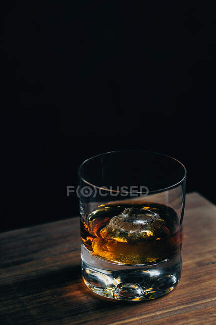 Сверху стеклянная чашка с холодным виски и кубиком льда, помещенная на деревянный стол в темной комнате — стоковое фото
