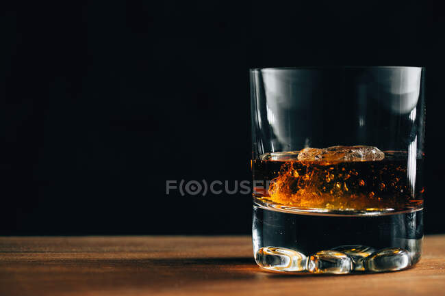 Copo de vidro com uísque frio e cubo de gelo colocado na mesa de madeira no quarto escuro — Fotografia de Stock