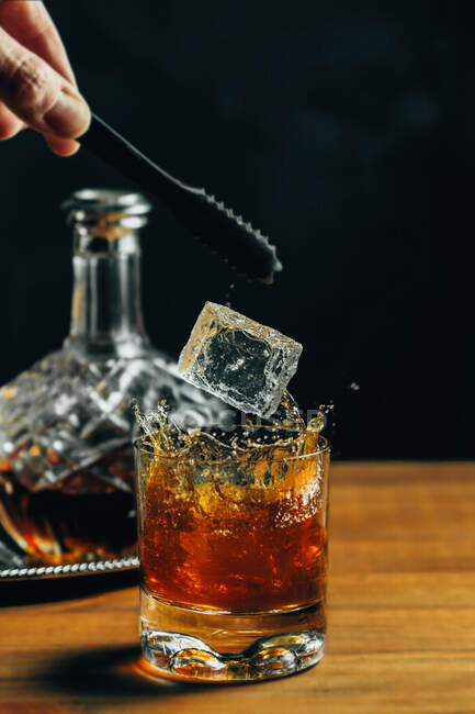 Vaso de whisky frío con hielo colocado sobre una mesa de madera cerca del decantador sobre fondo negro - foto de stock