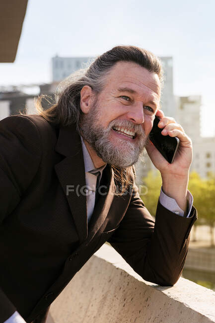 Бічний погляд на усміхненого дорослого чоловіка з сивим волоссям у класичному костюмі обговорюючи ділові проблеми під час телефонної розмови, стоячи навпроти сучасних міських будівель. — стокове фото