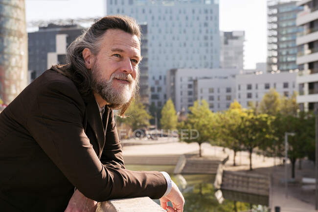 Side view of positive middle aged gray bearded bearded male in stylish formal outfit looking away pensively while leaning on fence on terrace of city building — стоковое фото