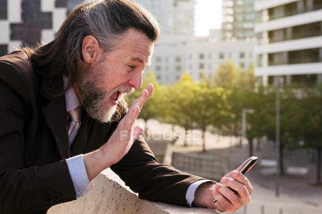 Вид сбоку на улыбающегося бородатого взрослого мужчину с седыми волосами в стильном костюме, размахивающего рукой во время приветствия партнера во время видеовстречи на смартфоне, стоя напротив современных городских зданий — стоковое фото