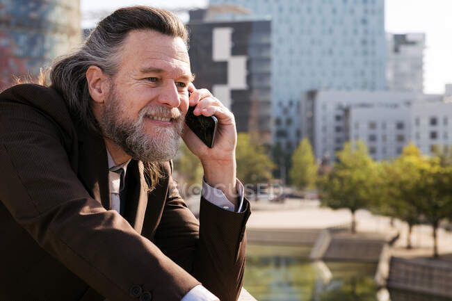 Vista lateral de hombre maduro barbudo sonriente con pelo gris que usa un traje elegante que discute temas de negocios durante la conversación telefónica mientras está parado contra los edificios modernos de la ciudad - foto de stock