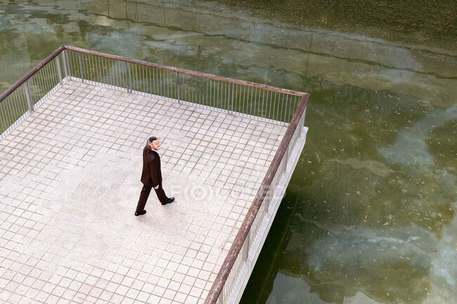 Desde arriba de solitaria elegante hombre de pelo gris en traje formal caminando solo en la plataforma vallada sobre el agua en la ciudad - foto de stock