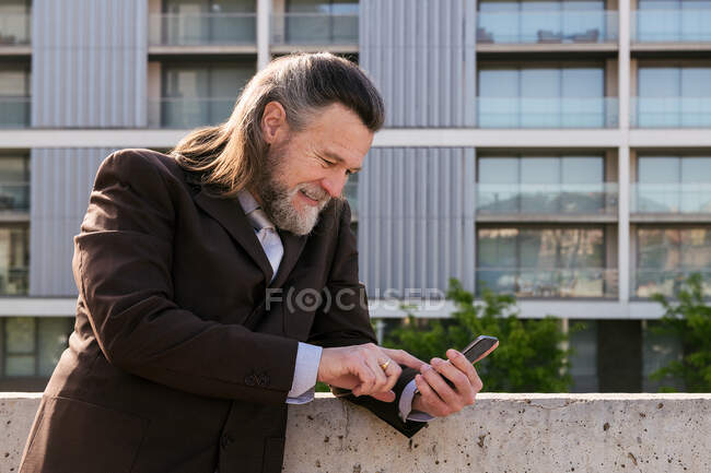 Side view of smiling bearded mature male with gray hair wearing classy suit browsing on smartphone while standing against modern city buildings — Stock Photo